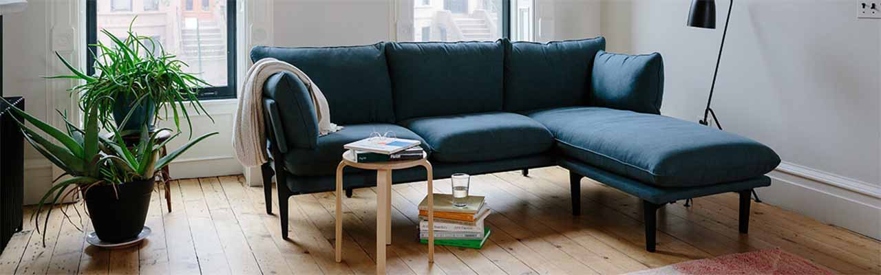 A modern living room with a dark blue sectional sofa, a light wooden coffee table with books and a glass of water, and a large green plant in a black pot on a wooden floor. The room has white walls, large windows with natural light, and a black floor lamp.
