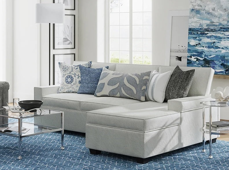 A modern living room featuring a light gray sectional sofa adorned with patterned and solid cushions. A glass coffee table with metal legs holds a black bowl and a book. The room has a blue patterned rug, large windows, and contemporary wall art and decor.