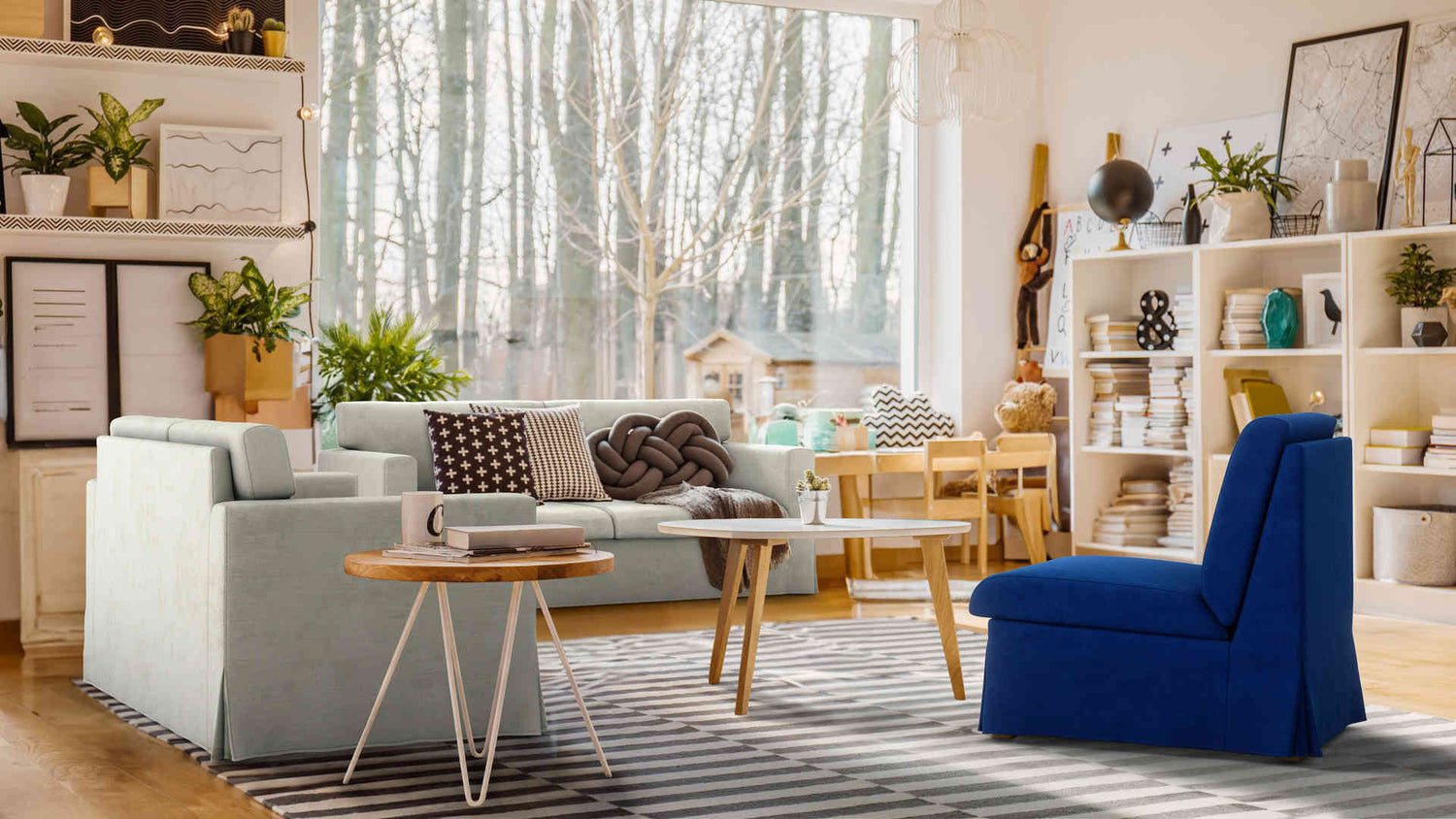 A cozy living room with large windows revealing a forest view. The room features a light blue sofa, a bold blue chair, and a textured rug. A wooden dining table with chairs is in the background, while shelves filled with books and plants adorn the space.