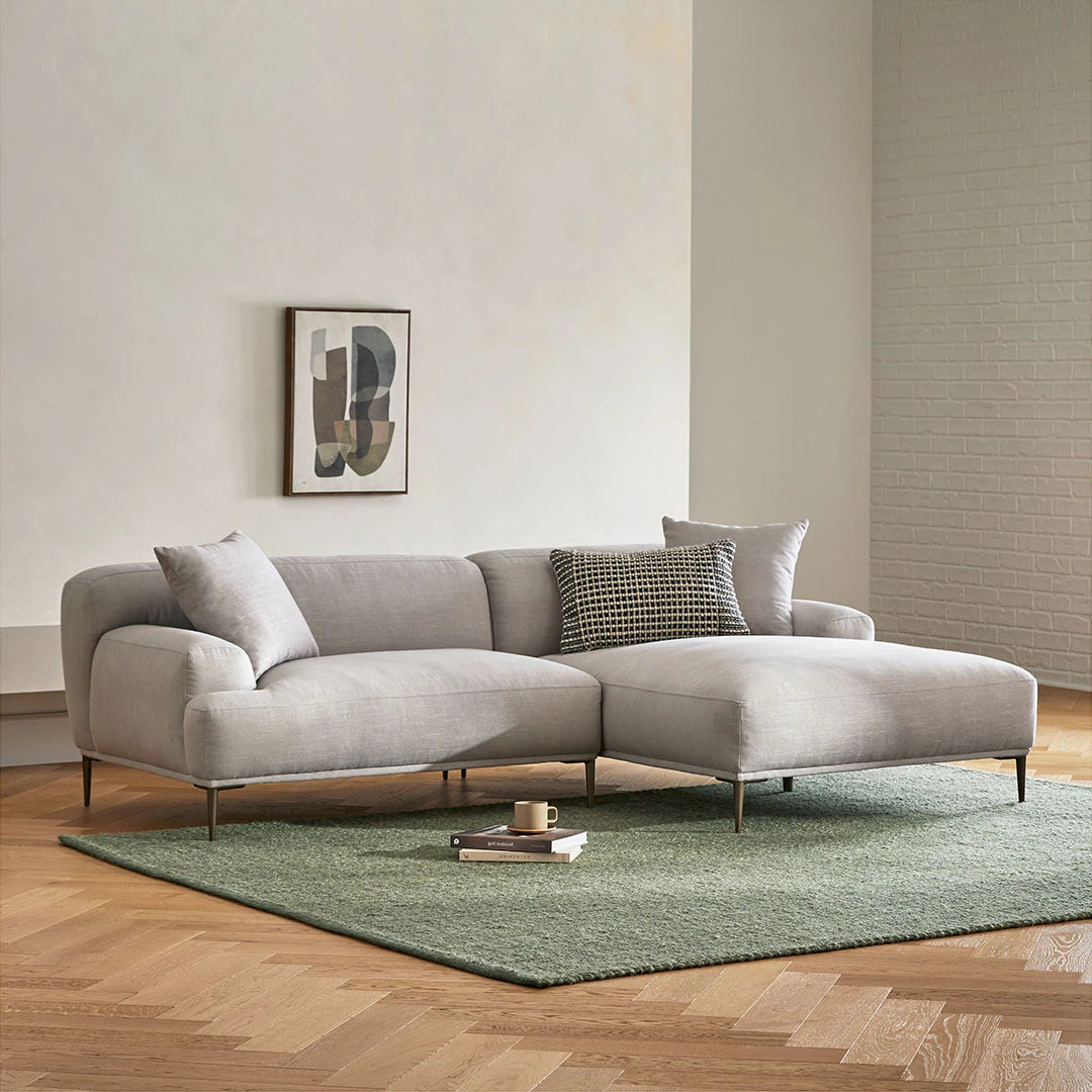 A modern living room showcases an Abisko 94.5 Right Sectional in Aurora Blue, accompanied by two cushions. It rests on a green area rug, with a small stack of books and a mug nearby. A minimalist abstract painting is displayed on the off-white wall, and the flooring is light wood.