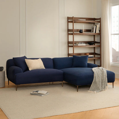 A cozy living room features the Abisko 94.5 Right Sectional in Aurora Blue, adorned with a cream pillow and a light gray throw blanket. A wooden shelving unit with decorative items rests against the wall, while soft natural light streams through a large window.