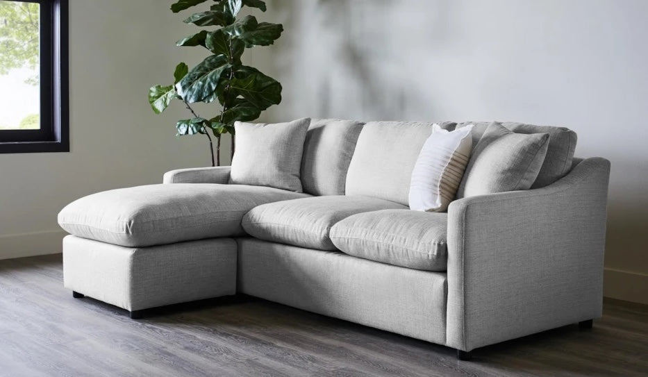 A modern living room features a light gray sectional sofa with a chaise lounge on the left side. A white and gray striped pillow is placed on the sofa. Behind the couch, a large green plant adds a touch of nature. The room has a minimalist design with wooden flooring.