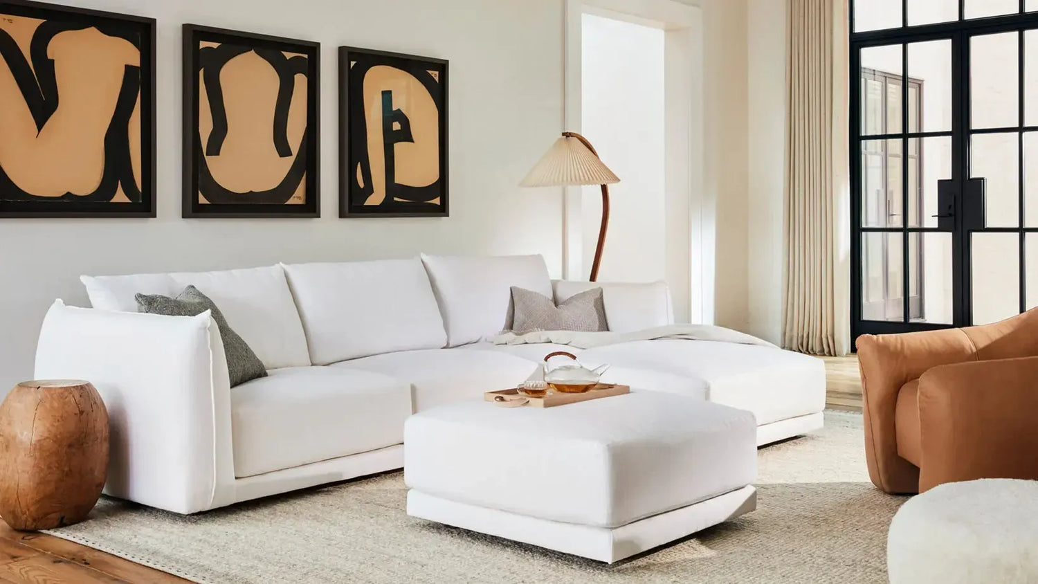A modern living room with a white sectional sofa, white ottoman, and a wooden side table. Three abstract art pieces hang on the wall behind the sofa. A standing lamp with a beige shade is placed next to the sofa. Large windows allow natural light to fill the space.