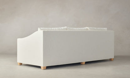 The Sullivan 60" Sofa in Performance Linen Weave, featuring a minimalist and modern design with a soft fabric cover in white and wooden legs, is set against a neutral gray background. The sofa's back is visible, highlighting its simple and clean aesthetic.