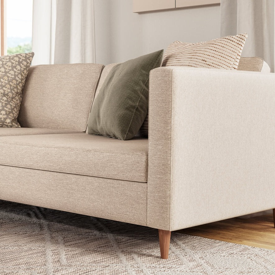 A beige sectional sofa with two green and one patterned throw pillows is positioned against a light-colored wall. The sofa is complemented by a beige area rug and sheer curtains, creating a cozy and neutral-toned living room setting.