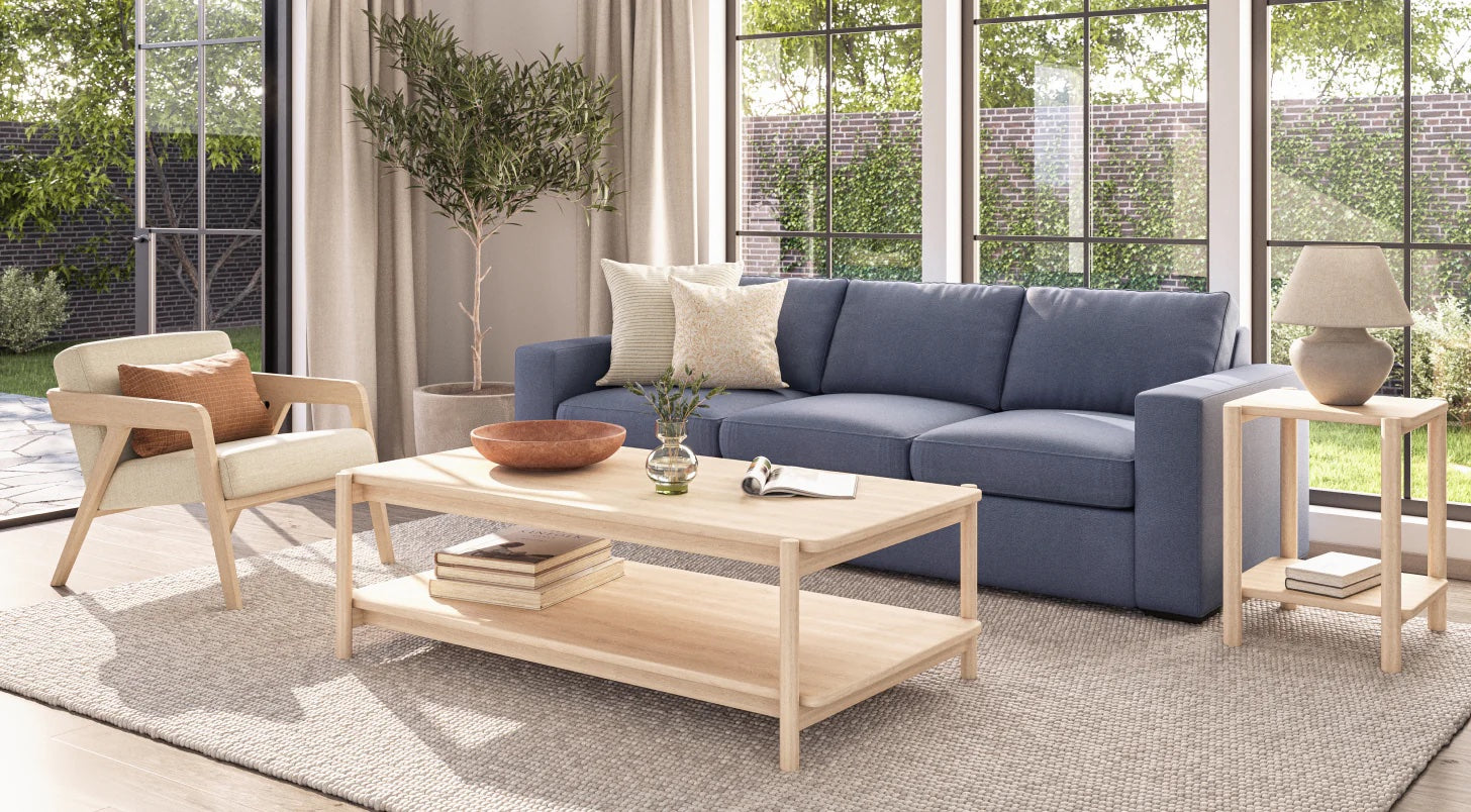 A bright living room with large windows overlooking a green garden. The room features a blue sofa with decorative pillows, a wooden coffee table with a vase and books, a beige armchair, a side table with a lamp, and a potted plant in the corner.