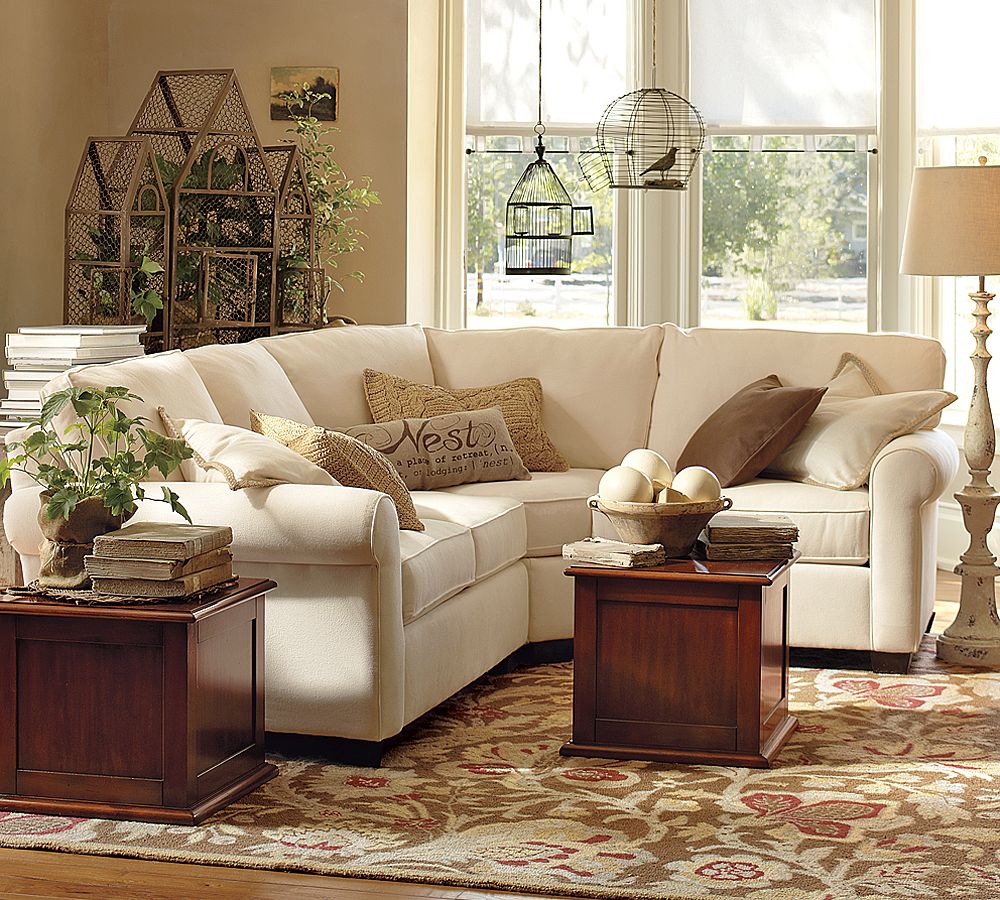 A cozy living room with a white sectional sofa adorned with brown and beige pillows. Two wooden end tables hold books and decorative items. Natural light streams through large windows, and the room features plants, a birdcage, and a patterned rug.