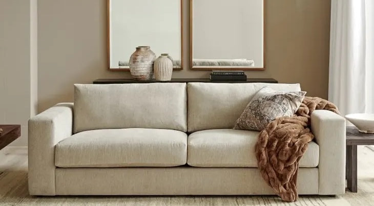 A cozy living room featuring a beige sofa adorned with a patterned pillow and a brown fur throw. Behind the sofa, there is a console table with decorative vases and books, and two large mirrors on the wall reflecting natural light from a nearby window.