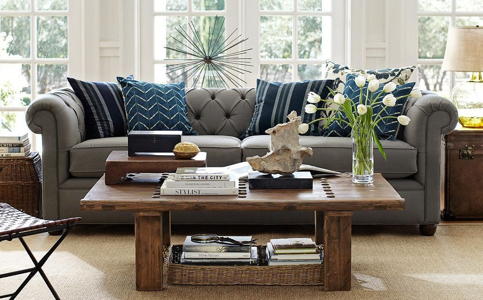 A cozy living room features a gray tufted sofa with blue and white patterned pillows. In front, a wooden coffee table is adorned with books, a decorative sculpture, and a vase of white tulips. Large windows in the background let in natural light.