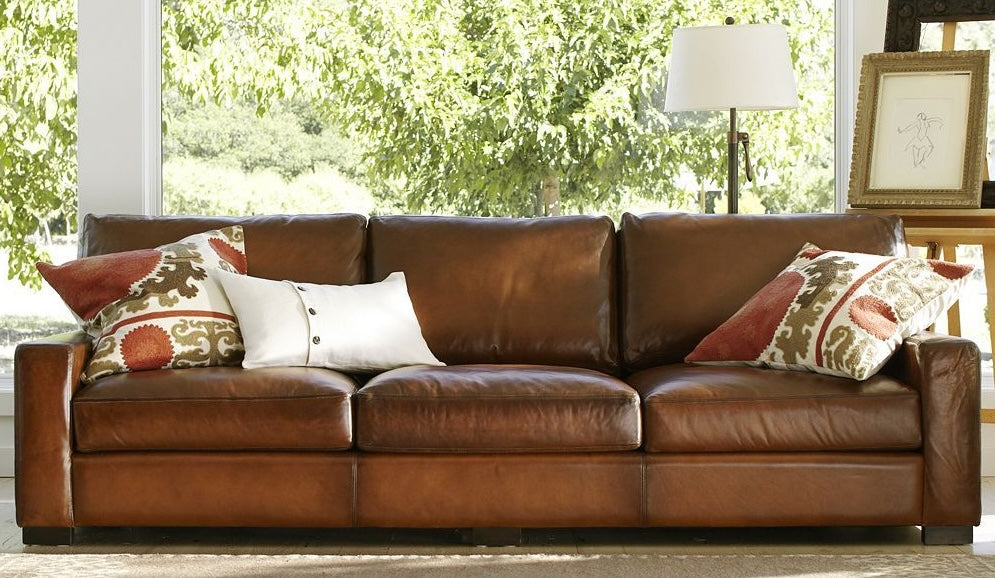 A cozy living room scene featuring a brown leather sofa adorned with three decorative pillows. Two pillows have a colorful pattern, while one is plain white with buttons. In the background, a tall window shows lush greenery, and a floor lamp and framed picture are to the right.