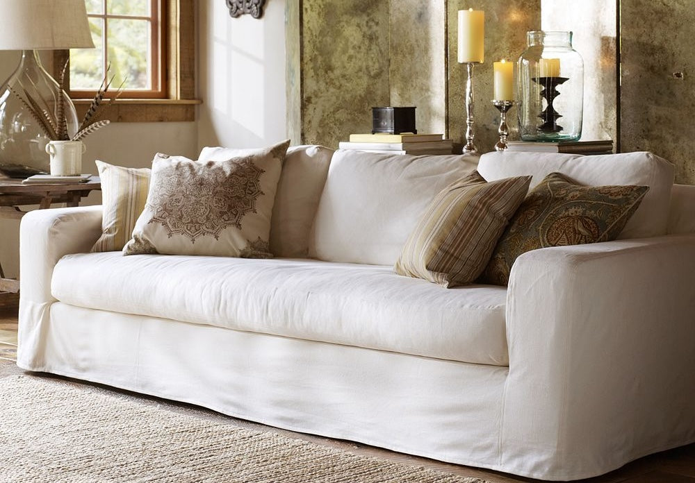 A cozy living room featuring a white sofa adorned with patterned throw pillows. Beside the sofa is a rustic wooden side table holding a lamp and decorative items. Candles, books, and other accessories are arranged on a shelf in the background. Natural light illuminates the space.