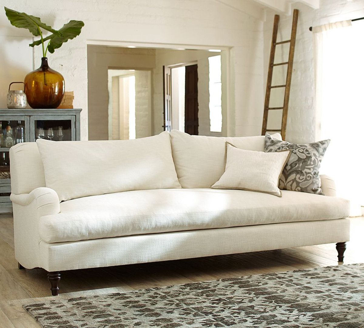 A cozy, well-lit living room features a large, off-white sofa adorned with matching and patterned throw pillows. Behind the sofa, there's a rustic cabinet with decorative items, including a large amber vase. A wooden ladder leans against the wall nearby.