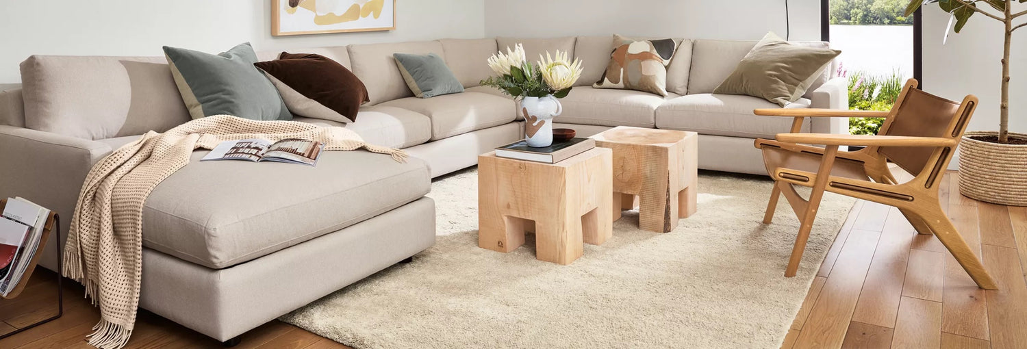 Modern living room featuring a large beige sofa with assorted cushions and a throw blanket, two natural wooden stools as a coffee table, a wooden armchair, and a cream rug over wooden flooring. A potted plant and abstract artwork add decorative touches.