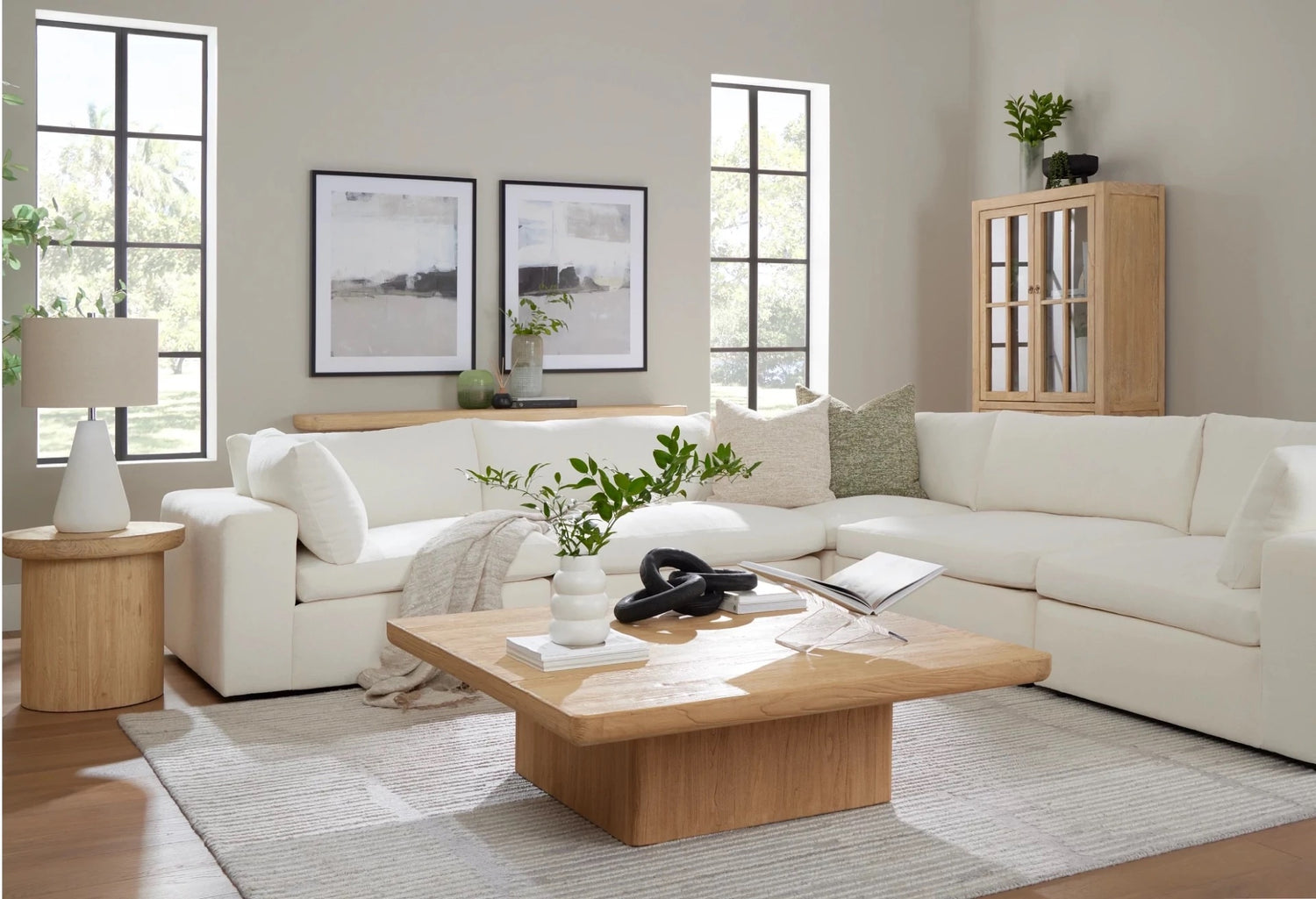 A contemporary living room features a white sectional sofa adorned with green and white throw pillows, a light wooden coffee table decorated with greenery and a book, matching side table with a lamp, and two abstract paintings on the wall. Large windows illuminate the space.
