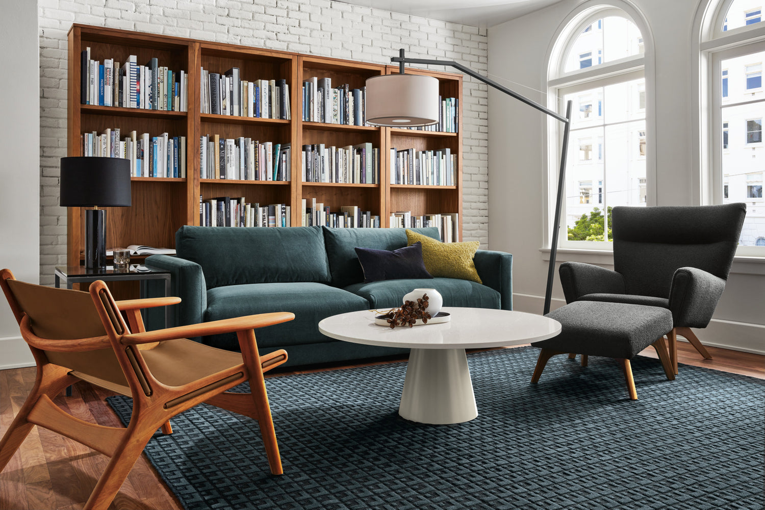 A modern living room features a teal velvet sofa with accent pillows, a white rounded coffee table, and two wooden chairs with cushioned seats. A tall arc floor lamp illuminates the space. Behind the sofa, a wooden bookshelf filled with books spans the wall. Large arched windows let in natural light.