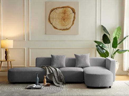 A modern living room features a grey sectional sofa with three cushions, a light grey throw draped on it, and two decorative vases on the floor. A large plant stands beside the sofa, and a wooden ring wall art hangs above. A lit lamp is in the left corner.