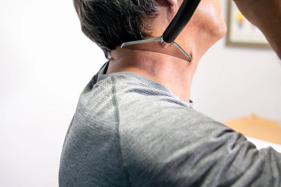 A person wearing a gray shirt holds a stethoscope to the back of their neck. The stethoscope's chest piece is resting on the skin, and the person's head is slightly tilted back. The background is out of focus with indistinct objects.