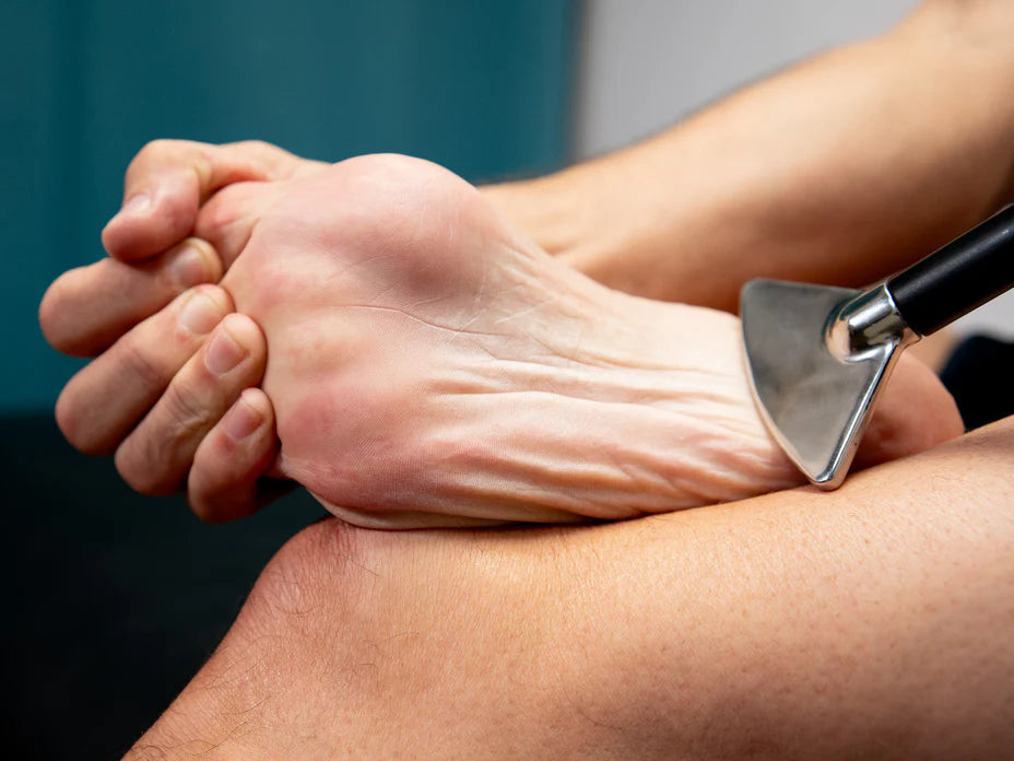A person is holding their foot with one hand, while another hand is using a metal tool to scrape the bottom of the foot, possibly for a massage or therapy. The background is blurred and out of focus.