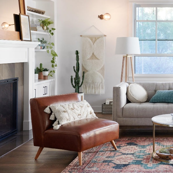 A cozy living room with a modern design features a brown leather chair with a textured pillow, a gray sofa with white and green pillows, a wooden coffee table, and a decorative rug. The room has a cactus plant, wall tapestry, floor lamp, and a large window.