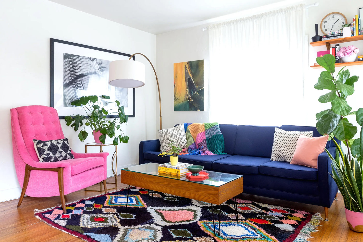 A vibrant living room featuring a blue sofa decorated with colorful pillows and a knitted throw. A pink armchair with a geometric cushion is beside a leafy plant. A wooden coffee table with books and plants sits on a patterned rug. Art and plants adorn the space.